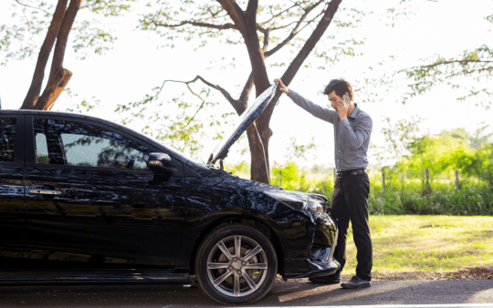 a man talking on a cell phone by a broken car on country road